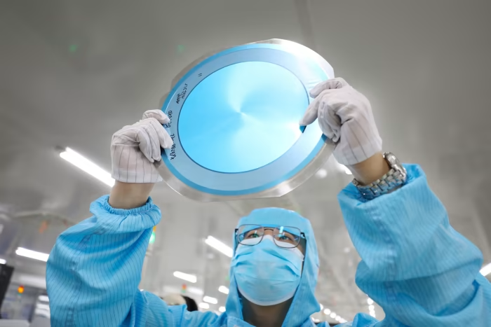 A worker in protective clothing, mask and gloves in a clean room, holds up a round disc to the light to look at electronic chips